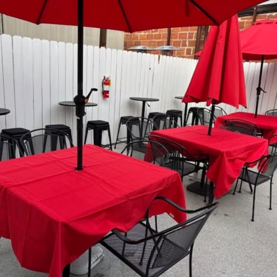 Outdoor patio with red-covered tables, black chairs, and red umbrellas against a white fence.