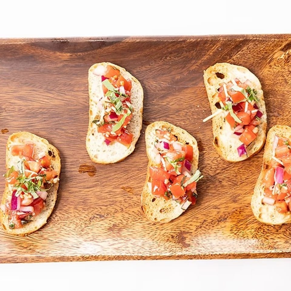 Five bruschetta pieces with diced tomatoes, red onion, and basil on a wooden serving board.