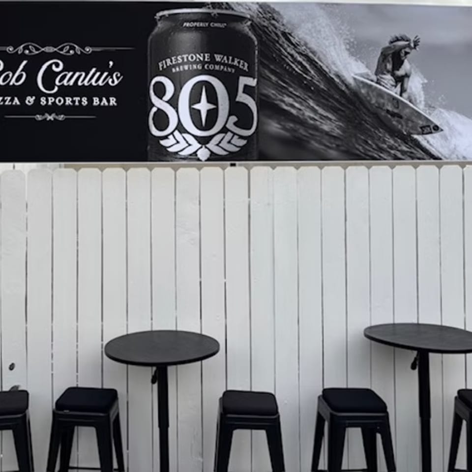 Outdoor seating area with black tables and stools against a white fence, below a Bob Cantu's sign.