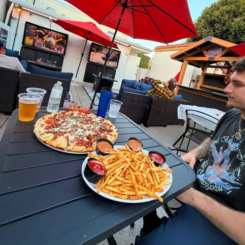Pizza and fries on an outdoor patio table with people, red umbrellas, and TVs in the background.