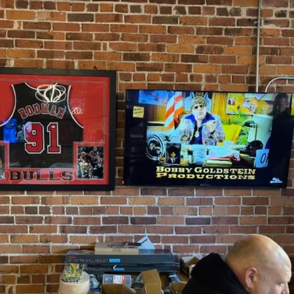 A framed Rodman Bulls jersey and a TV playing "Bobby Goldstein Productions" against a brick wall.