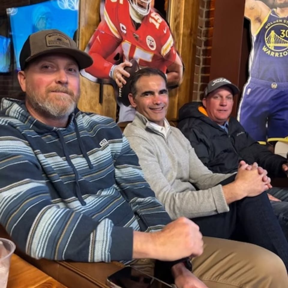 Three men sitting on a couch in a sports bar, with sports cutouts in the background.