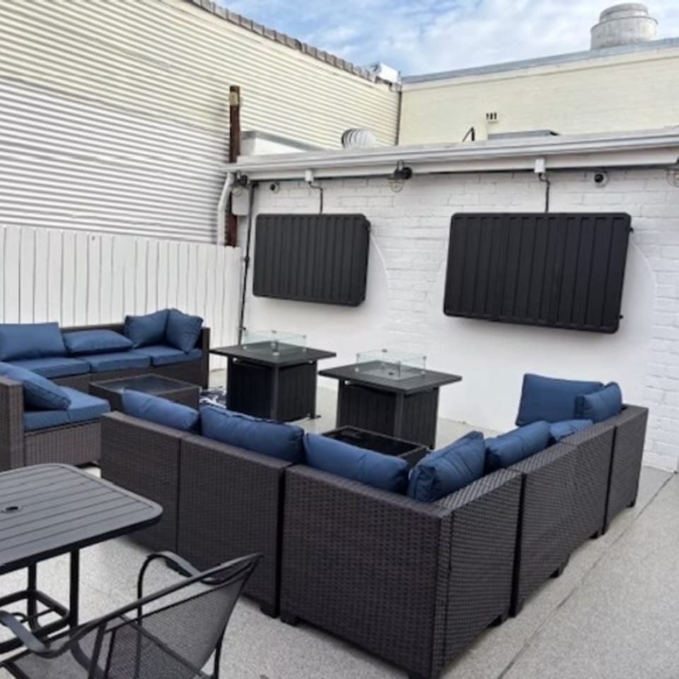 Outdoor patio seating area with dark wicker sofas, blue cushions, fire pit tables, and wall-mounted TVs.